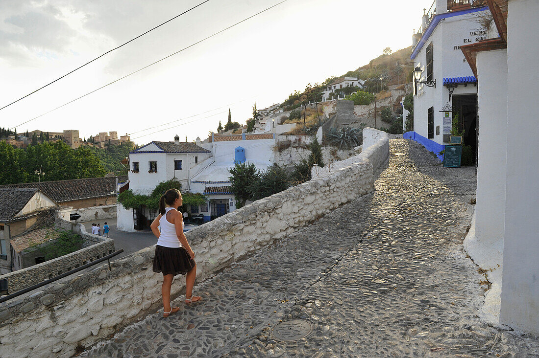Girl walking uphill on a cobblestone lane in Sacromonte with the Alhambra in the background, Granada, Andalusia, Spain