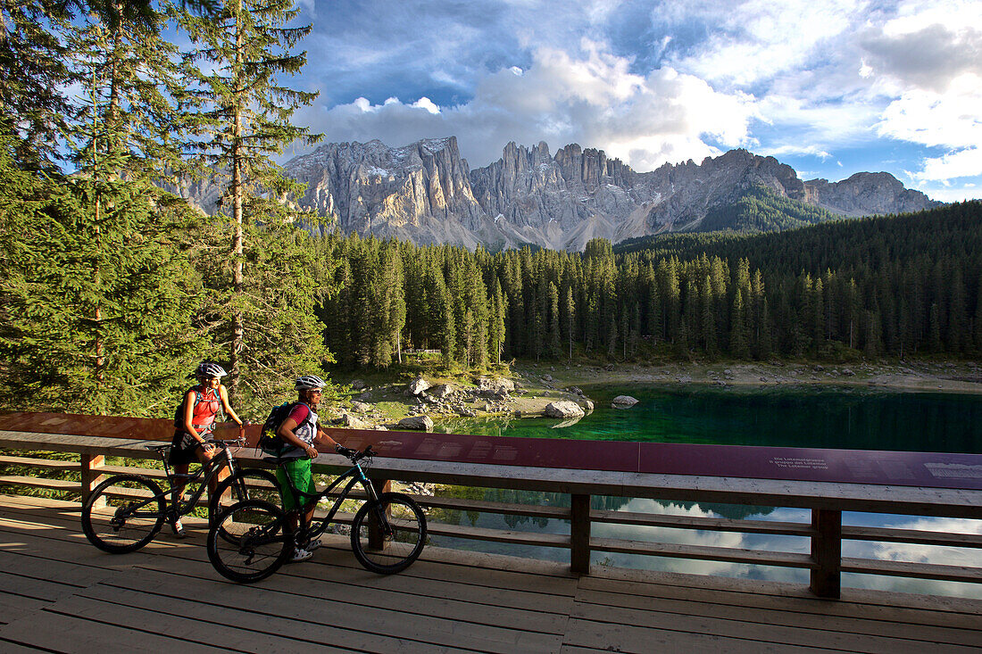 two mountain bikers on an observation platform at lake Karersee, Latemar massif in the background, Trentino, Italy