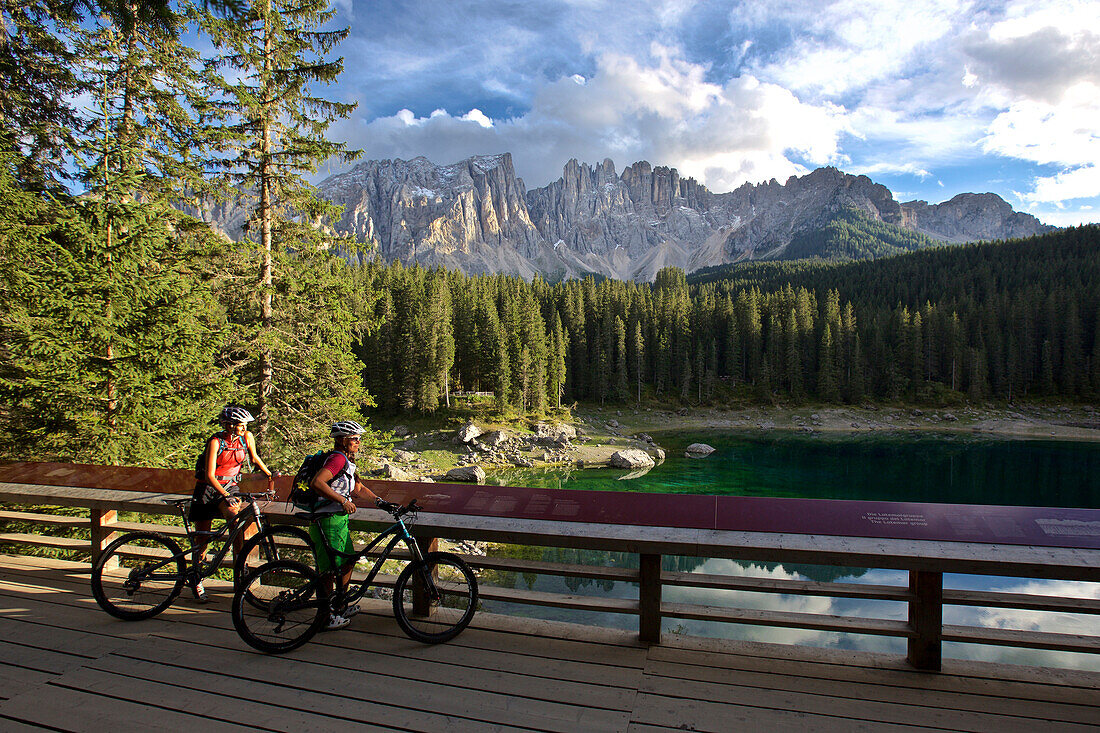 zwei Mountainbikerinnen auf einer Aussichtsplattform am Karersee, dahinter Latemarmassiv, Trentino, Italien