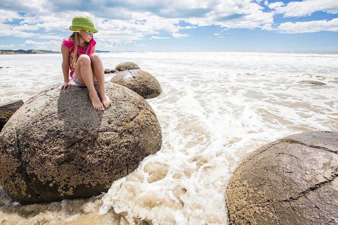 A girl sitting on a Moreaki Boulder with the rising tide, Moeraki Boulders, South Island, New Zealand