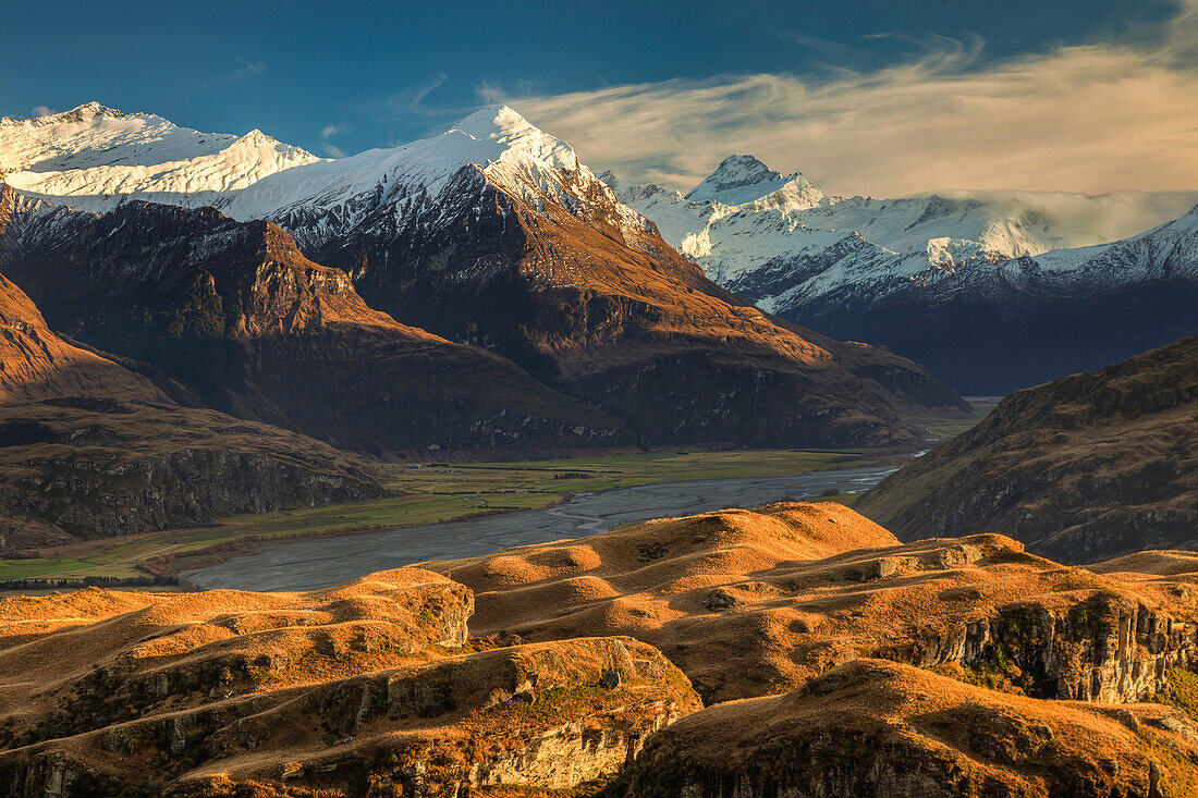 Dawn light across tussock grass hills above Matukituki River and Black Peak and Mount Aspiring near Wanaka, Otago, New Zealand