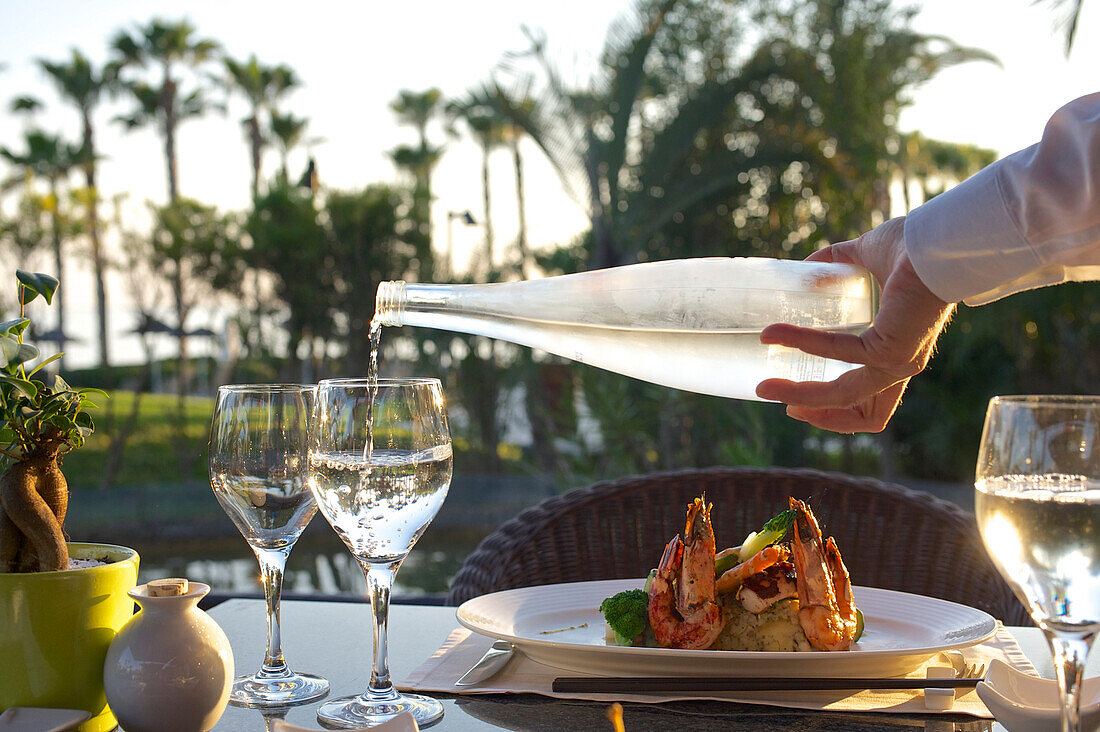 Gedeck mit Gläsern, Riesengarnelen und Hand, die Wasser einschenkt im Gartenrestaurant mit Abendsonne und Palmen im Hintergrund im Le Meridien Hotel, Limassol, Limassol District, Zypern