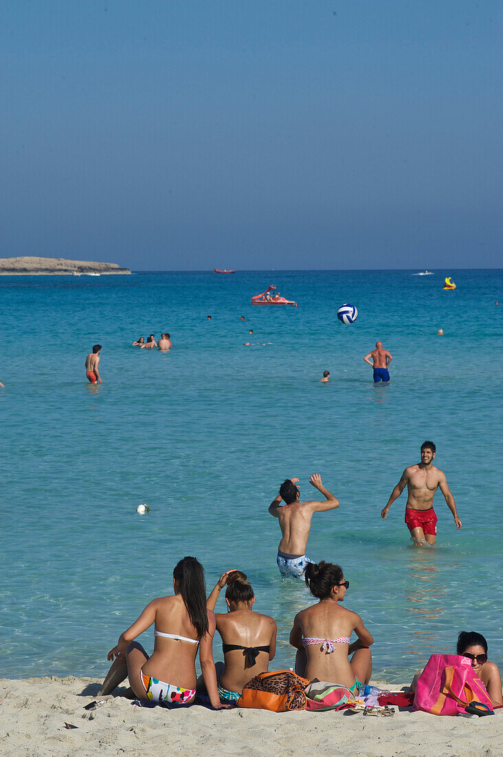 White sandy beach with three women sitting on the beach and men playing volleyball in the water, Nissi Beach near Ayia Napa northeast of Larnaca, Larnaca District, Cyprus