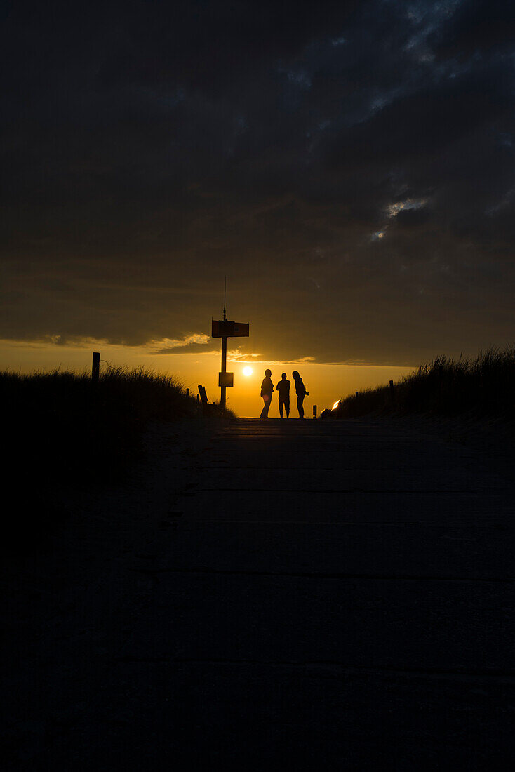 Drei Personen stehen am Strandaufgang und unterhalten sich im Gegenlicht des Sonnenuntergangs , Nationalpark Vorpommersche Boddenlandschaft, Dierhagen, Mecklenburg Vorpommern, Deutschland