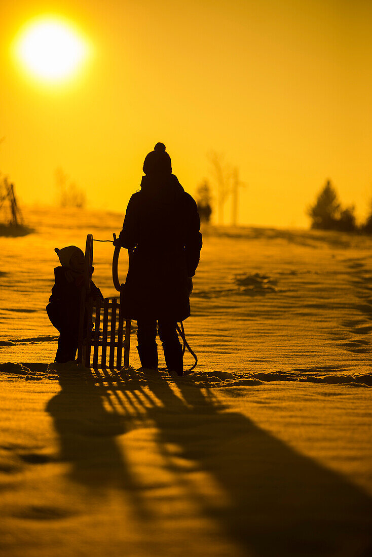 Mutter steht mit kleinem Kind und Schlitten im Schnee und schauen die Wintersonne an, Gegenlichtaufnahme, Aubing, München, Bayern, Deutschland