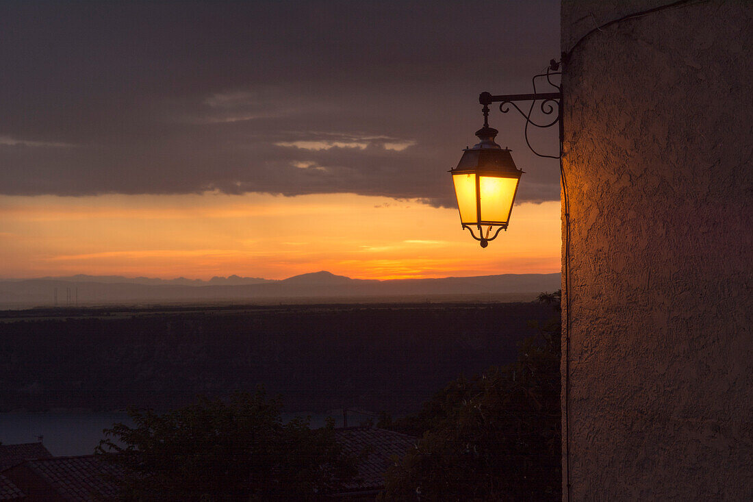 A street lamp glowing, with the setting sun in the background, village of Aiguines, Var department, Provence-Alpes-Cote d’Azur region, France