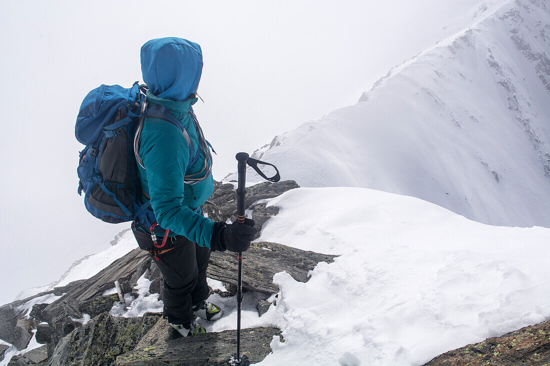 Eine Alpinistin in Skitourenausrüstung steht auf einem exponierten Grat im kombinierten Gelände und blickt hinab auf ihre Spur am Anfang des Grats, Wasenhorn respektive Punta Terrarossa, Simplongebiet, Lepontinische Alpen, Kanton Wallis, Schweiz