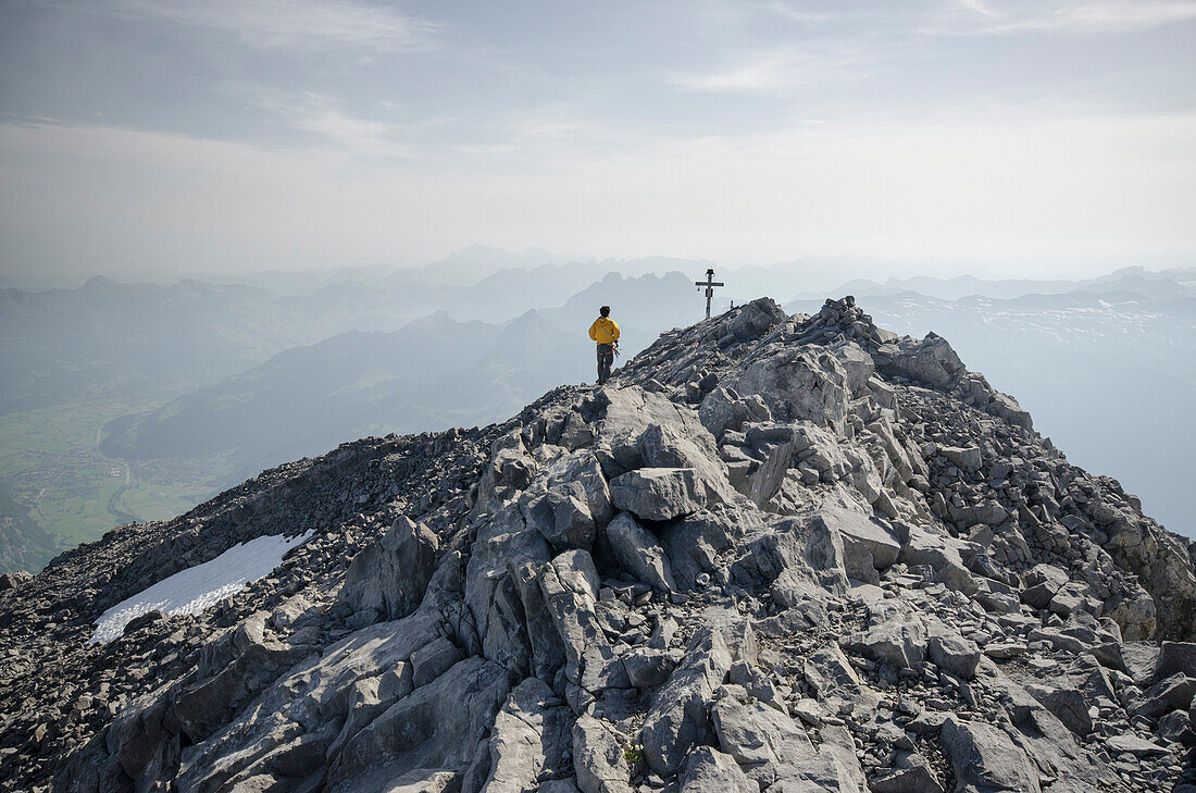 An alpinist standing next to the summit cross on the summit of Vrenelisgaertli, on the left in the valley the villages of Naefels and Mollis, Glarus Alps, canton of Glarus, Switzerland
