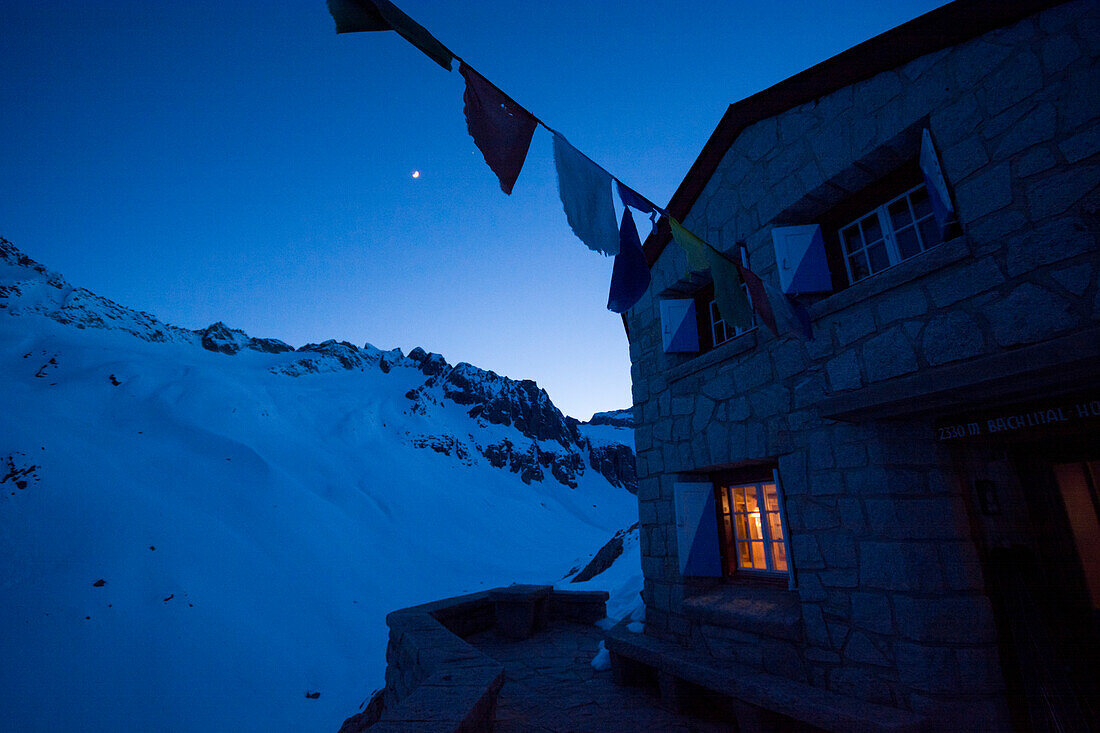 Im Stubenfenster der Bächlitalhütte brennt Licht, während es draußen dämmert und der Mond scheint, Bächlital, Grimselgebiet, Berner Alpen, Kanton Bern, Schweiz