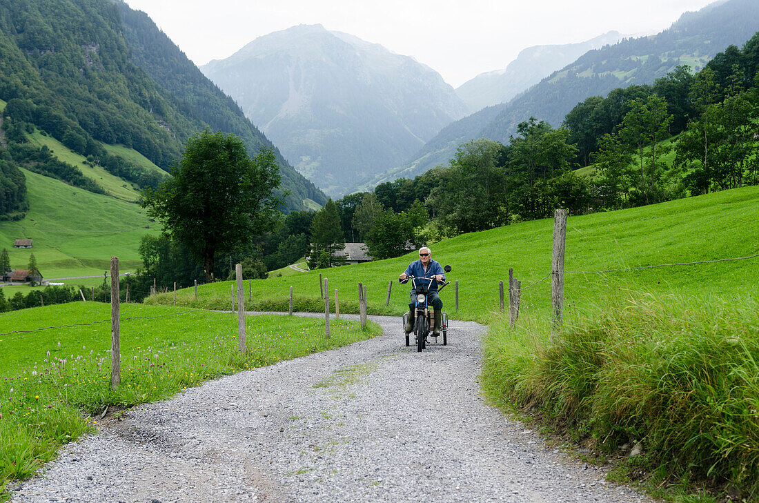 Ein älterer Mann fährt auf einem Moped über einen Kiesweg, Sernftal, Kanton Glarus, Schweiz