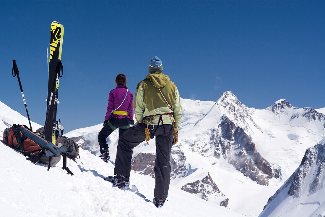 Eine Bergsteigerin und ein Bergsteiger mit Skitourenausrüstung stehen auf dem Gipfel der Roccia Nera, Breithorn-Massiv, im Hintergrund der Gipfel der Dufourspitze, Monte-Rosa-Massiv, Walliser Alpen, Kanton Wallis und Region Aostatal, Landesgrenze Schweiz 