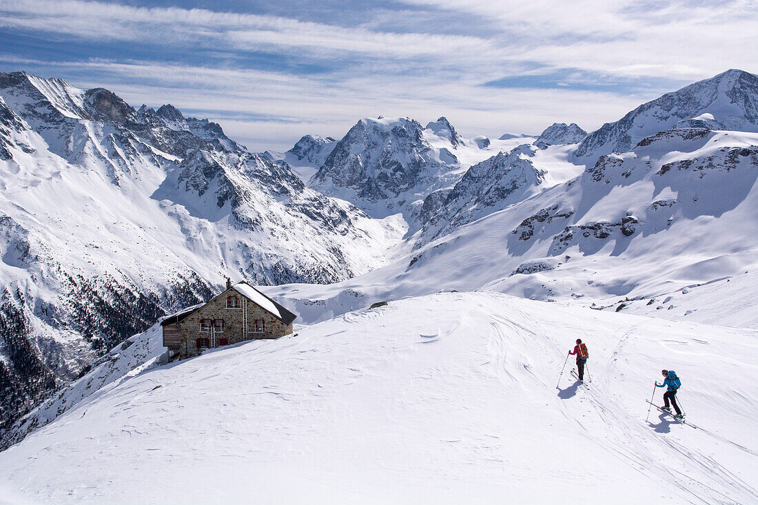 Two female backcountry skiers reaching the Aiguilles Rouges Hut above the village of Arolla over a small, snow-covered plateau, behind the summits of Mont Collon in the centre and Pigne d‘Arolla on the far right, Val d‘Herens, Pennine Alps, canton of Vala