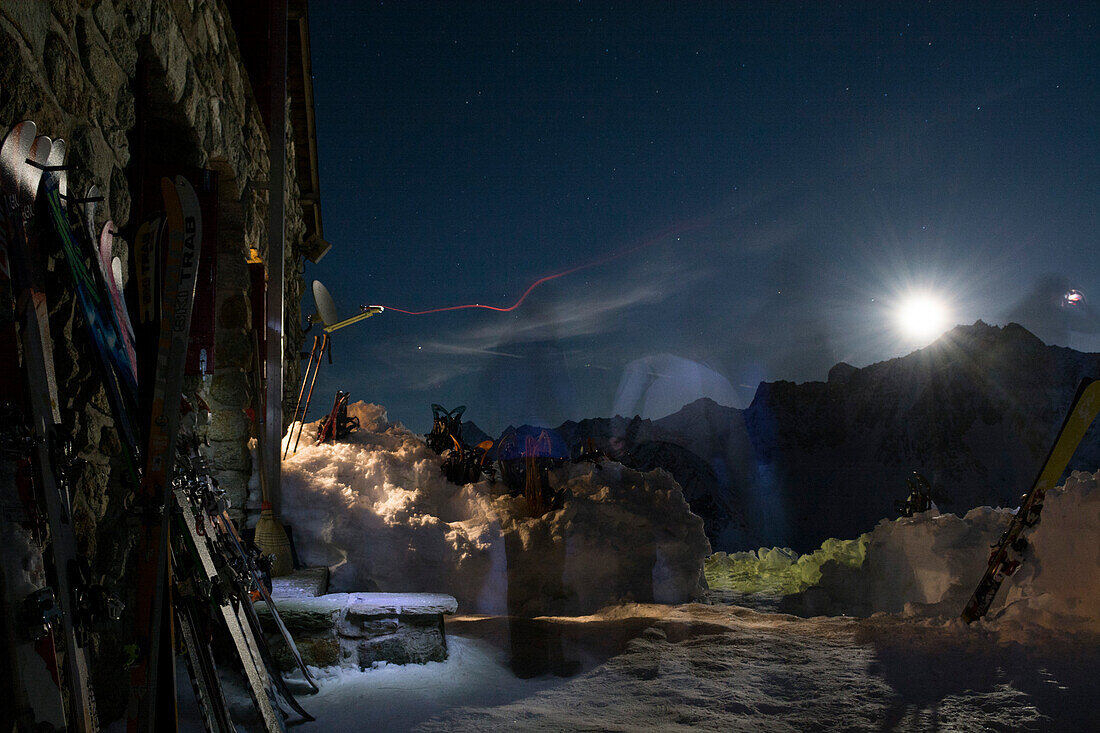 Nachts vor der Cabane des Aiguilles Rouges bewegen sich Gäste schemenhaft mit Stirnlampen im Dunkeln, der Mond scheint derweil hell über eine Bergkette und beleuchtet die Steinfassade und die daran angelehnten Ski, Val d‘Hérens, Walliser Alpen, Kanton Wal