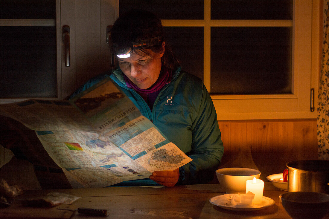 Eine Frau mit Stirnlampe sitzt am Tisch bei Kerzenlicht und studiert die Skitourenkarte,  Cadlimo Hütte, Lepontinische Alpen, Kanton Tessin, Schweiz