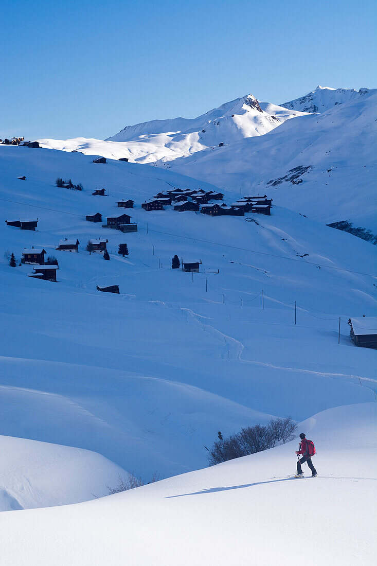 A backcountry skier walking through the snow-covered high valley called Fondei, behind him the houses of the hamlet Strassberg, Grison Alps, canton of Grison, Switzerland