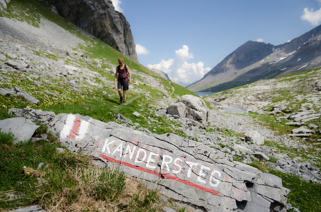 Eine junge Frau wandert auf einem Wanderweg, im Vordergrund an einem Stein eine Wanderwegmarkierung, die den Weg ins Dorf Kandersteg weist, im Hinergrund der Daubensee, Wanderung über den Pass namens Gemmi, Berner Alpen, Kantone Bern und Wallis, Schweiz
