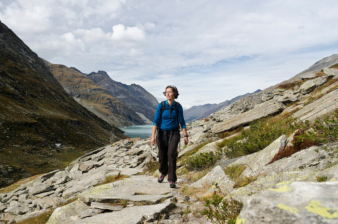 A young female hiker on a stone path hiking towards Monte Moro Pass, behind her Mattmark reservoir, Pennine Alps, canton of Valais, Switzerland