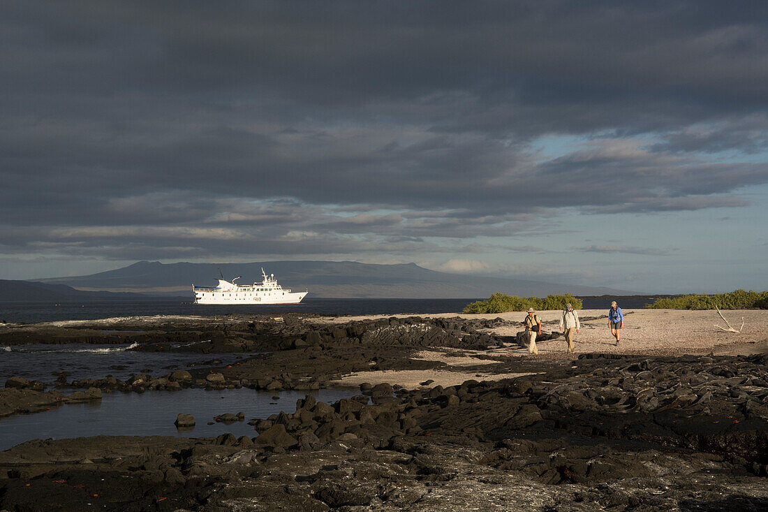 Ein Führer und zwei Touristen wandern über Sand und schwarze Vulkanfelsen bei Punta Espinoza auf der Insel Fernandina, im Hintergrund vor Anker das Boot La Pinta, Galapagosinseln, Ecuador