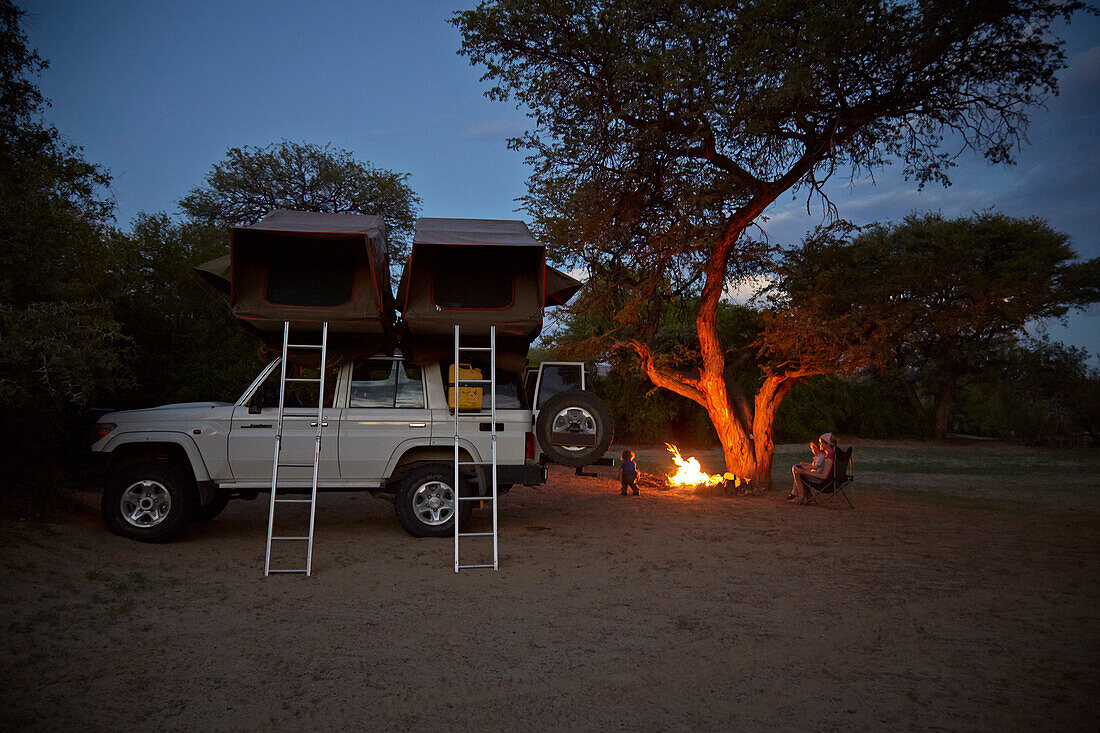 Family around a campfire, off-road vehicle with roof top tent in foreground, Purros Camp, Hoarusib, Namib desert, Nambia