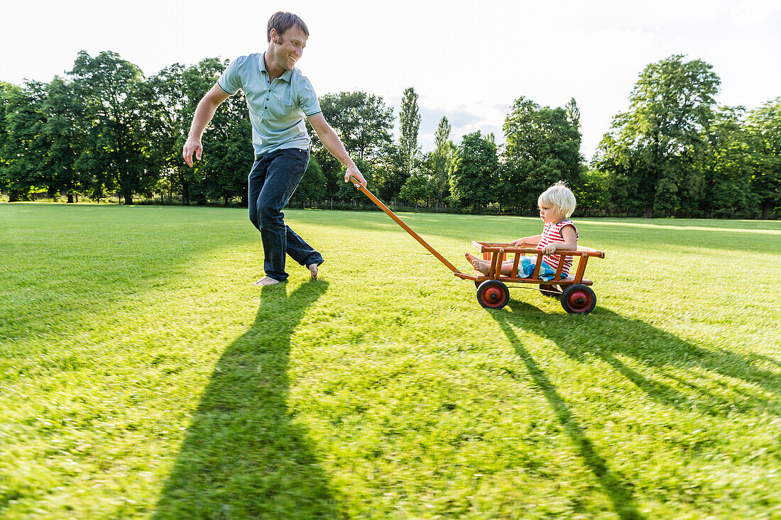 young father pulling his two-year-old daughter in a wagon over a meadow, Speyer, Rheinland-Pfalz, Germany