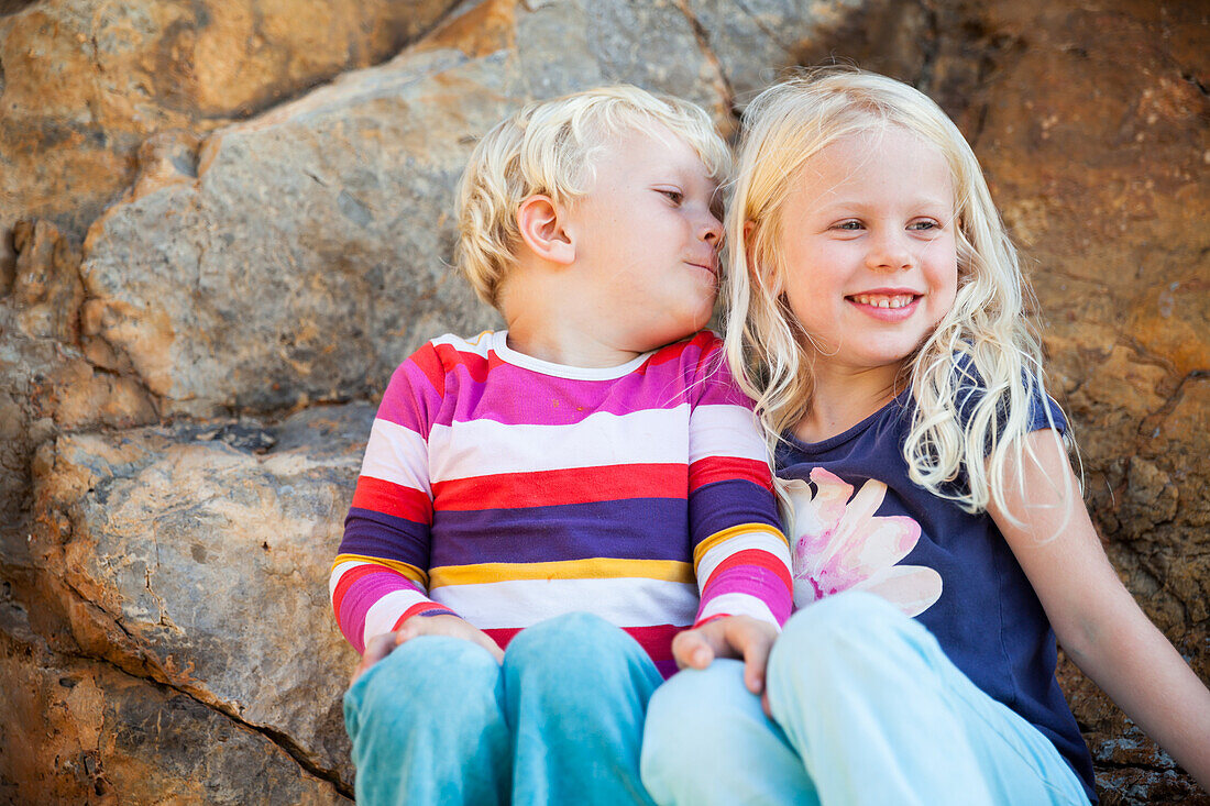 Junge und Mädchen auf Felsen, sitzen gemeinsam am Felsen und lachen, Port de Soller, Mallorca, Balearen, Spanien, Europa