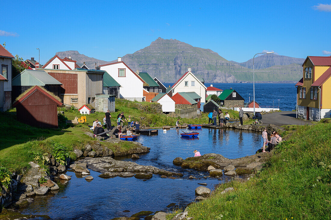 Kinder spielen auf dem Dorfteich zwischen den bunten Häusern von Gjogv und vor der Fjordlandschaft, Insel Eysturoy, Färöer Inseln (Føroyar)