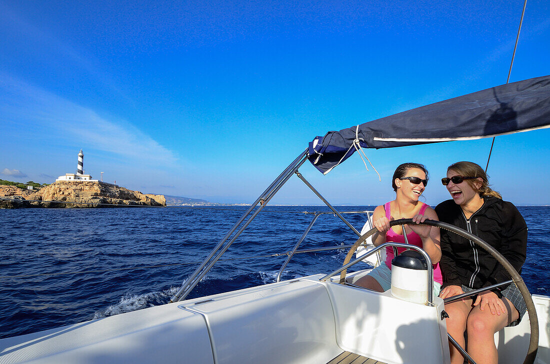 Zwei junge Frauen sitzen am Steuerrad einer Segelyacht, steuern und passieren das Cap Figuera mit dem Leuchtturm Faro Cala Figuera, im Hintergrund Palma de Mallorca, Balearen, Spanien, Europa