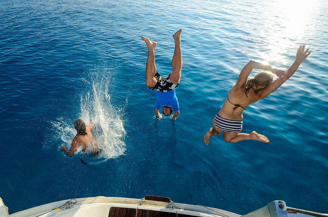 Two young women and a young man are jumping from the stern of a sailing yacht into the clear blue sea, Mallorca, Balearic Islands, Spain, Europe