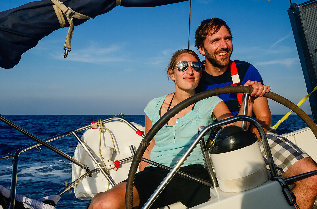 A young woman sitting at the steering wheel, rudder of a sailing yacht, a young man is sitting behind her, Mallorca, Balearic Islands, Spain, Europe