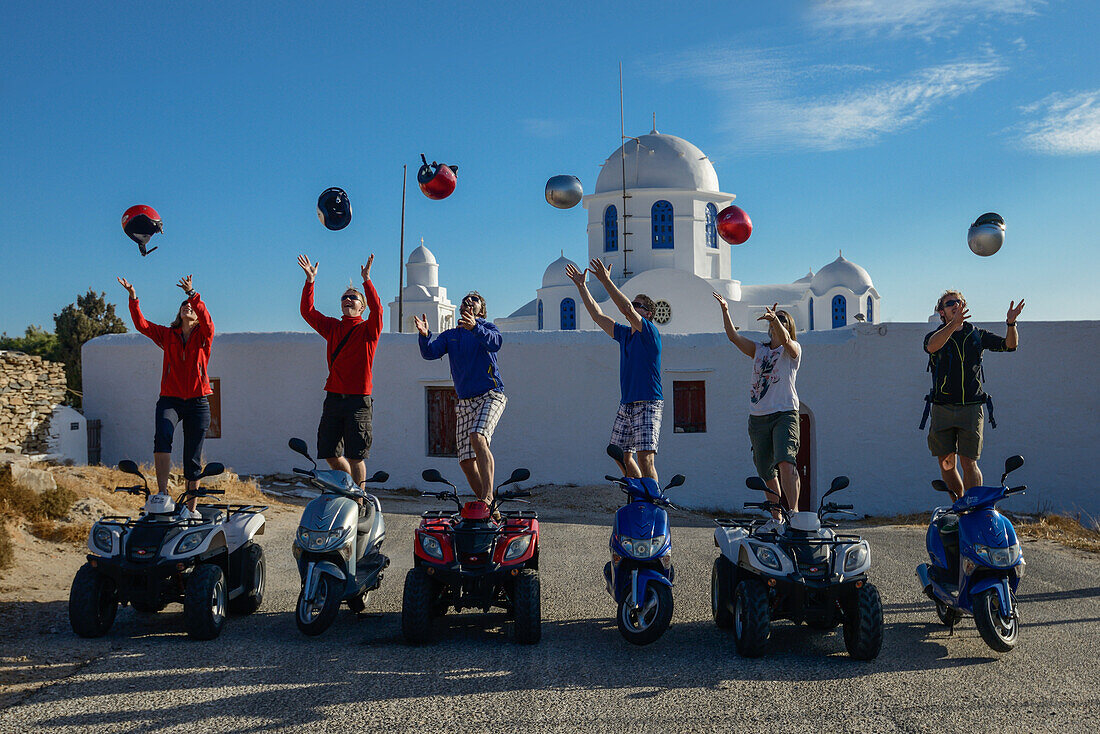 Drei junge Frauen und Männer stehen auf Motorrollern und Quads (Quad bikes) und werfen ihre Helme in die Luft, Sifnos Kirche Eftamartyres auf der griechischen  Insel Sifnos (Syphnos), Ägäis, Kykladen, Griechenland