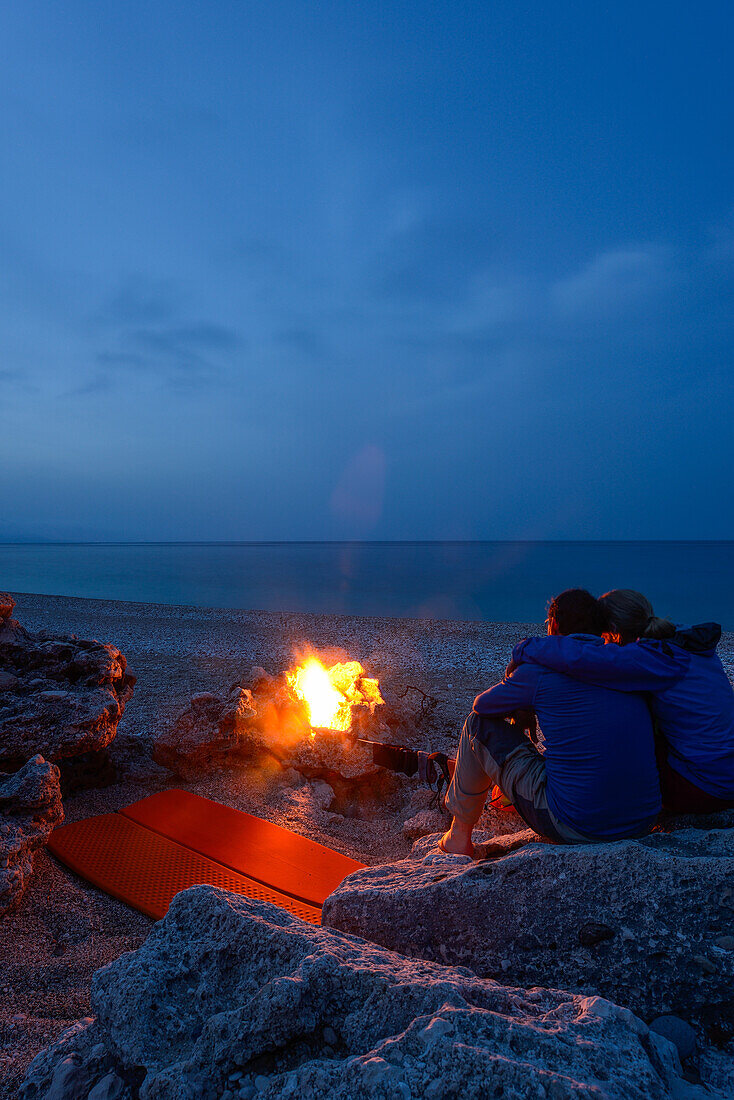 Young woman and young man sitting on rocks near the camp fire and camping out on the pebble beach of the bay Cala Sisine, Golfo di Orosei, Selvaggio Blu, Sardinia, Italy, Europe
