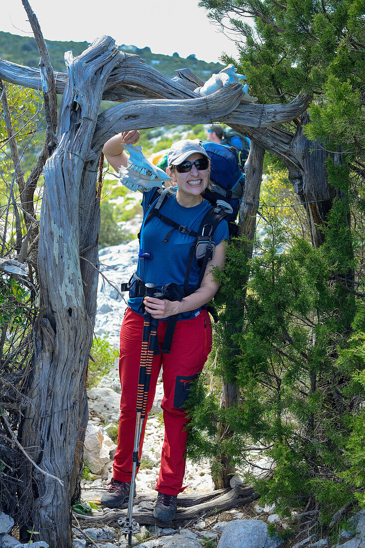 Junge Frau mit Trekkingausrüstung geht durch ein Tor aus Wacholderstämmen und hält einen Tierschädel in der Hand, Selvaggio Blu, Sardinien, Italien, Europa