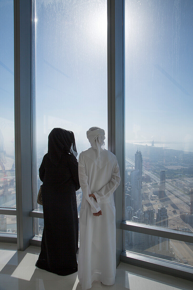 Frau und Mann in arabischer Kleidung blicken Blicken auf Skyline von Aussichtsplattform At The Top im 124. Stockwerk vom Burj Khalifa Turm, Dubai, Vereinigte Arabische Emirate