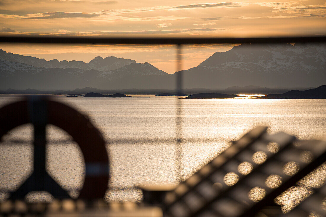 Silhouette of deck chairs and life ring aboard cruise ship MS Deutschland (Reederei Peter Deilmann) at sunset with mountains in distance, near Lofoten, Norway