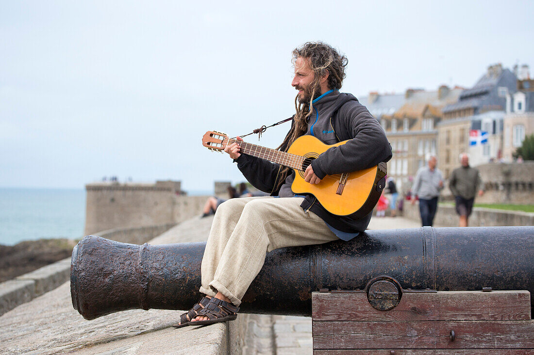 Straßenmusiker sitzt auf Kanone auf der Stadtmauer der alten Festung und spielt Gitarre, Saint-Malo, Bretagne, Frankreich, Europa