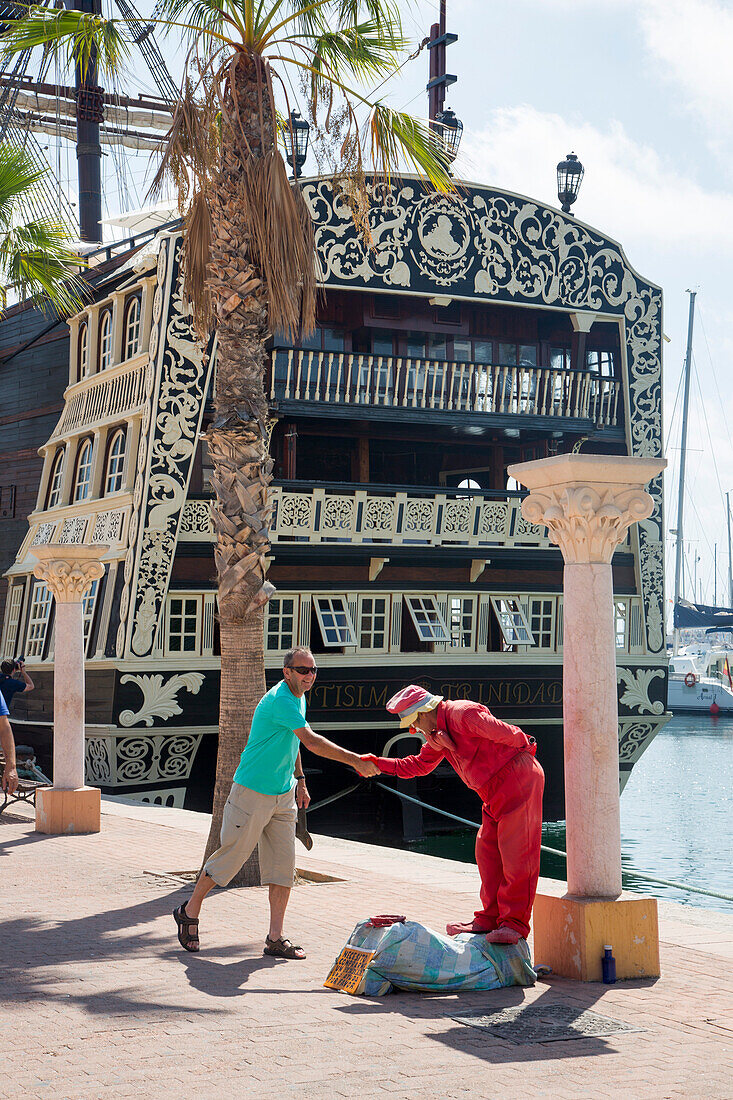 Clown Street performer shakes hands with man with replica of historic sailing ship Santisima Trinidad in port behind, Alicante, Andalusia, Spain