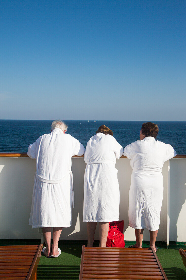 Drei ältere Passagiere in weißen Bademänteln an der Reling von Kreuzfahrtschiff MS Deutschland (Reederei Peter Deilmann) im Mittelmeer, nahe Spanien, Europa