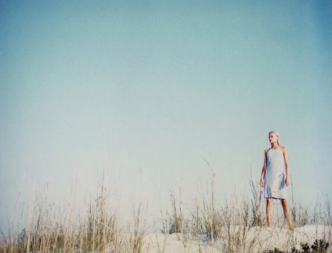 Woman standing on grassy sand dune at beach, staring at distance