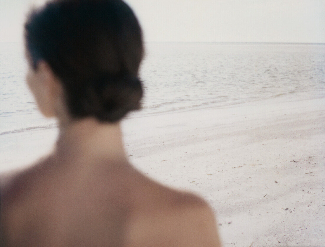 Woman looking toward ocean, rear view of head and shoulders, focus of beach in background