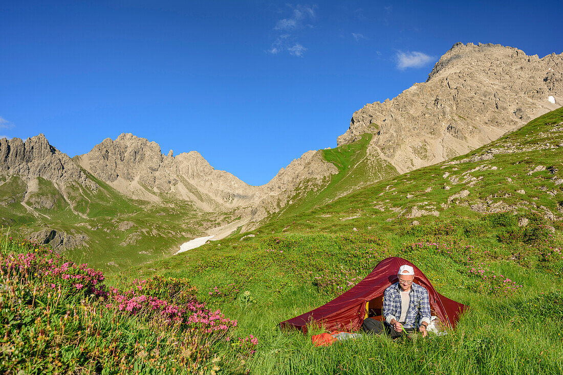 Mann sitzt vor Zelt in Wiese mit Alpenrosen, Steinkarspitze im Hintergrund, Lechtaler Alpen, Tirol, Österreich