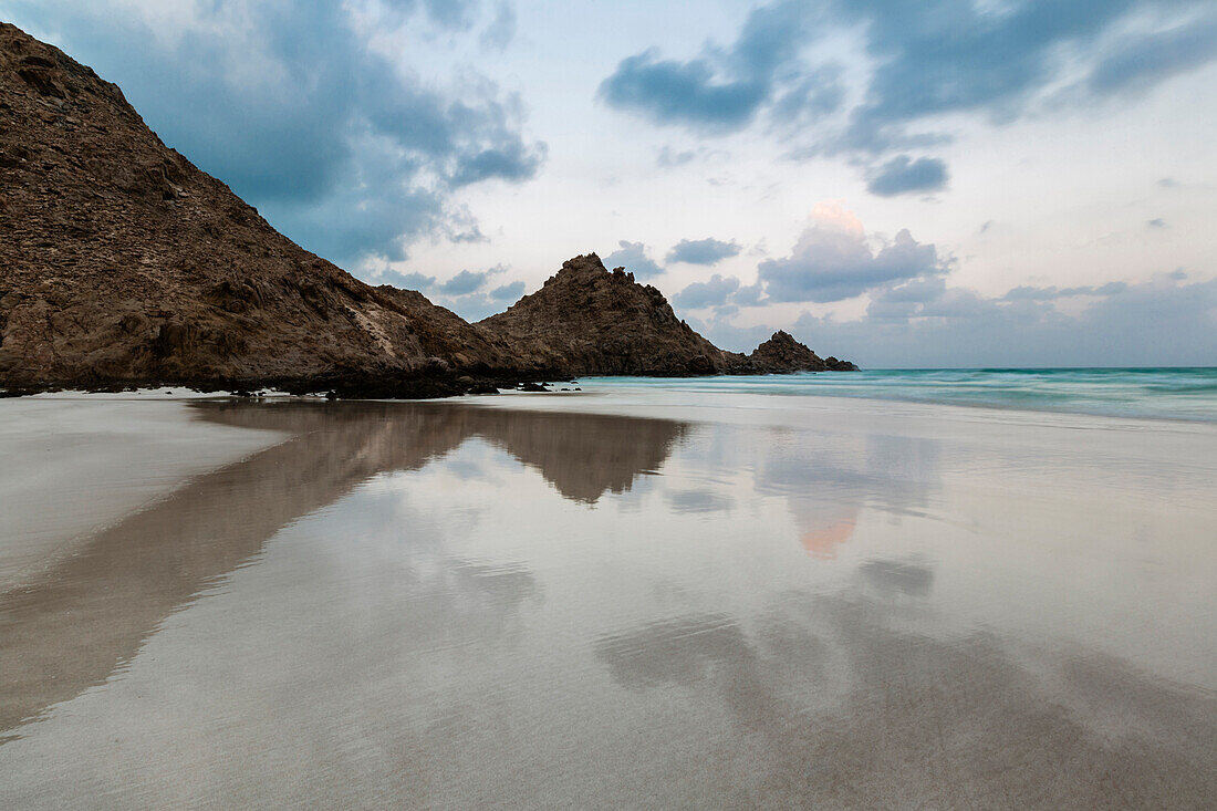 Rocky coastline reflected in still beach surf, Qalansyia, Socotra, Yemen