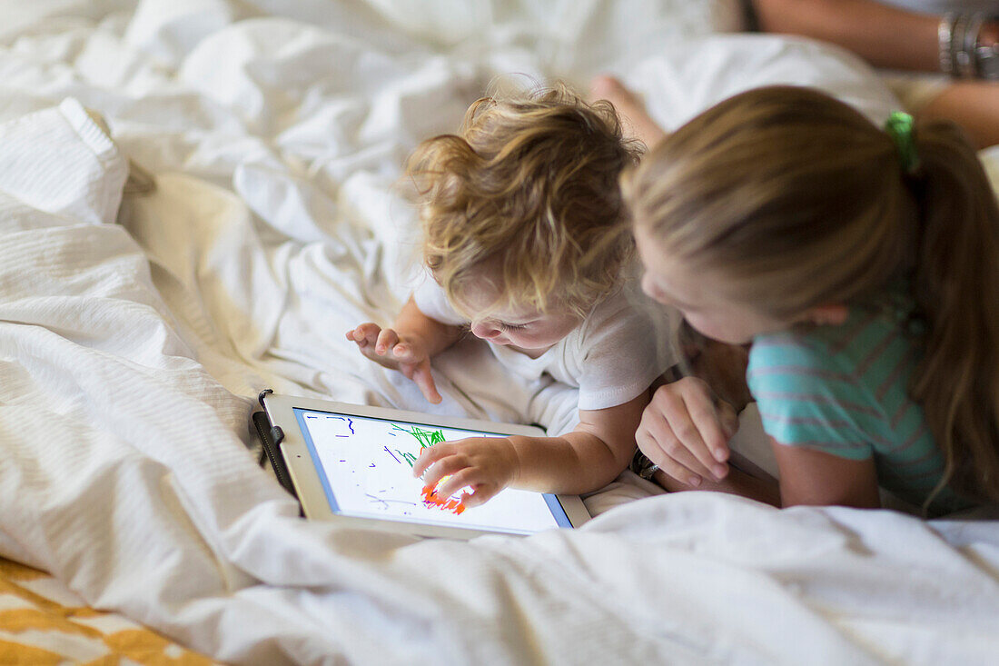 Caucasian girl and toddler brother using tablet computer on bed, Santa Fe, New Mexico, USA