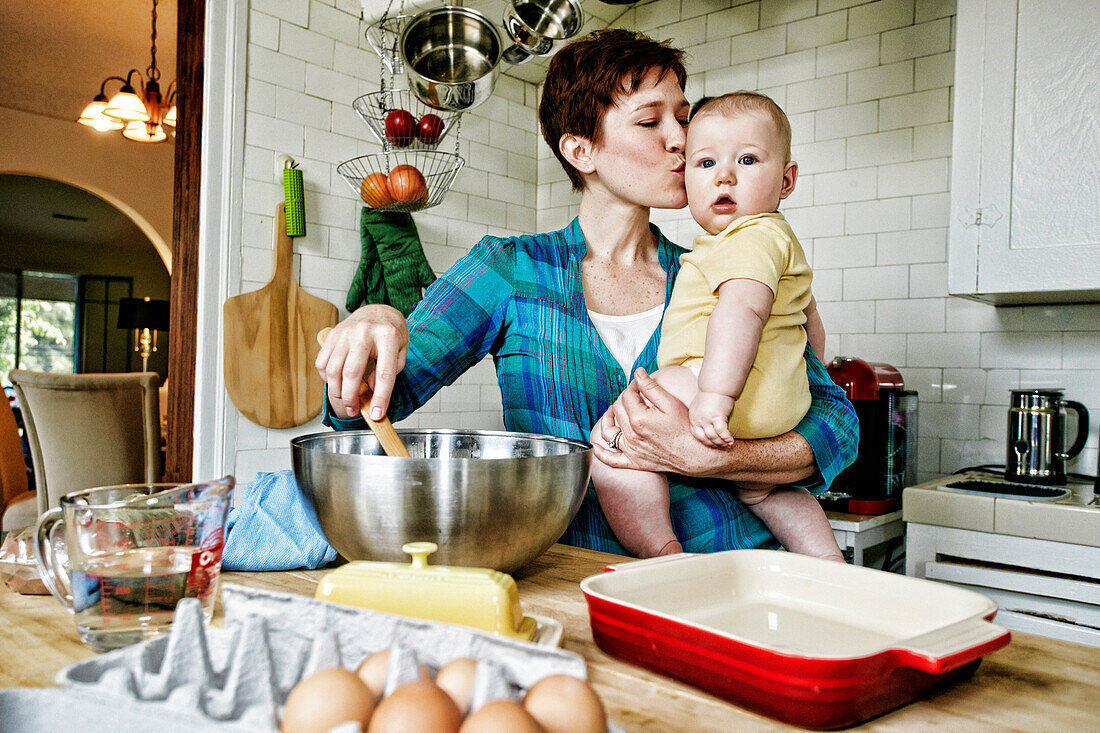 Caucasian mother kissing baby and cooking in kitchen, C1