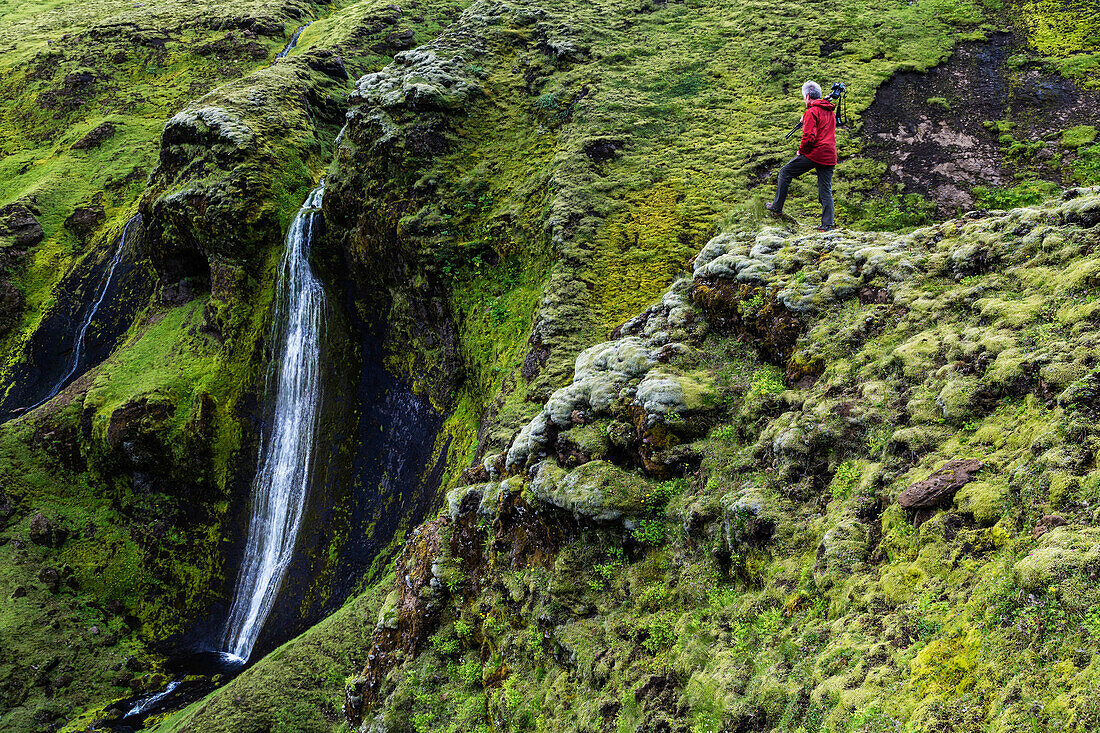 Caucasian hiker admiring waterfall and rock formations on mountainside, Southern Highlands, Iceland, ICELAND