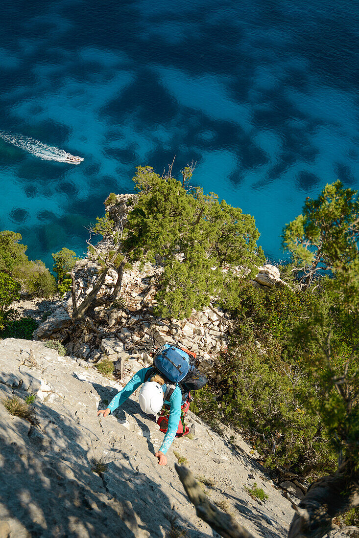 A young woman climbing a steep wall above the sea, while a motor yacht passes, Golfo di Orosei, Selvaggio Blu, Sardinia, Italy, Europe