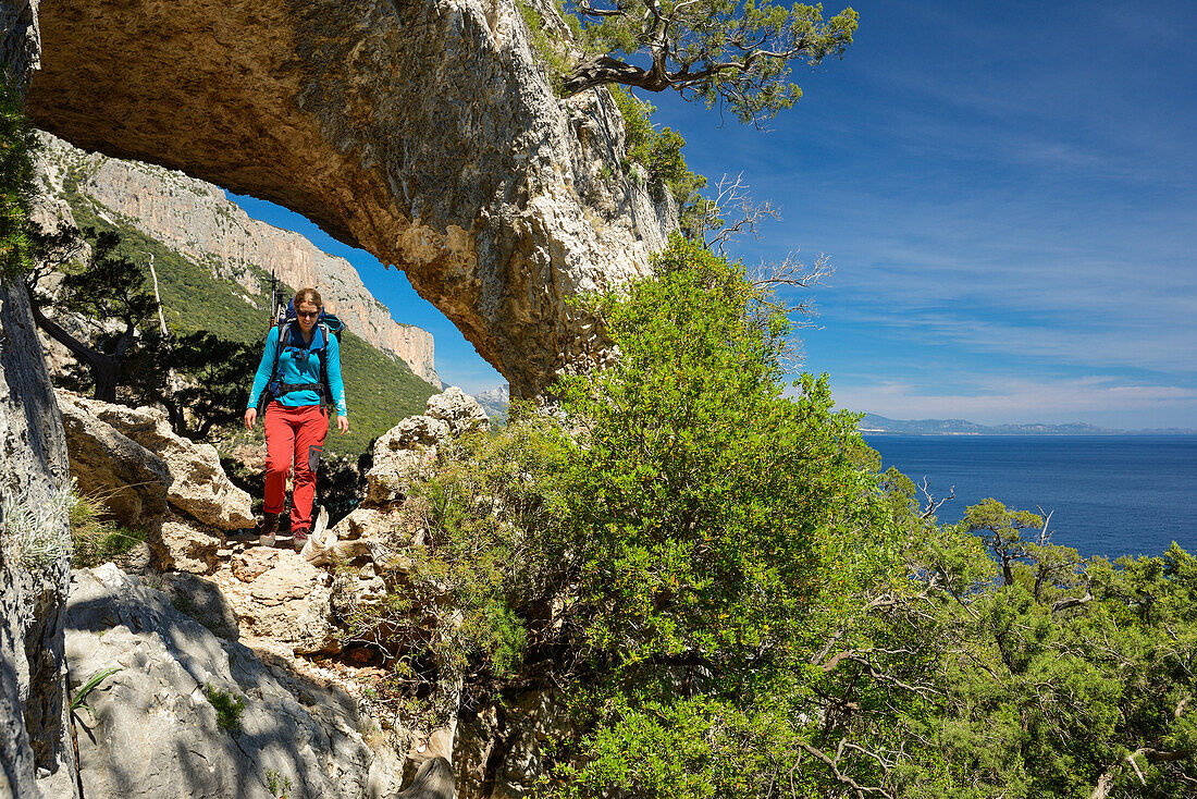 Eine junge Frau mit Trekkingausrüstung wandert durch den Felsbogen Arcu su Feilau oberhalb des Meeres entlang der gebirgigen Küste, Golfo di Orosei, Selvaggio Blu, Sardinien, Italien, Europa