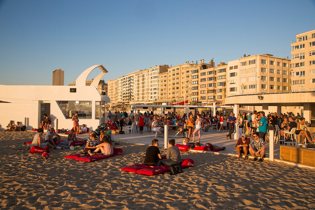 Junge Menschen chillen an einer Strandbar am Strand mit Apartments an der Strandpromenade im Hintergrund, Ostende, Flandern, Belgien, Europa