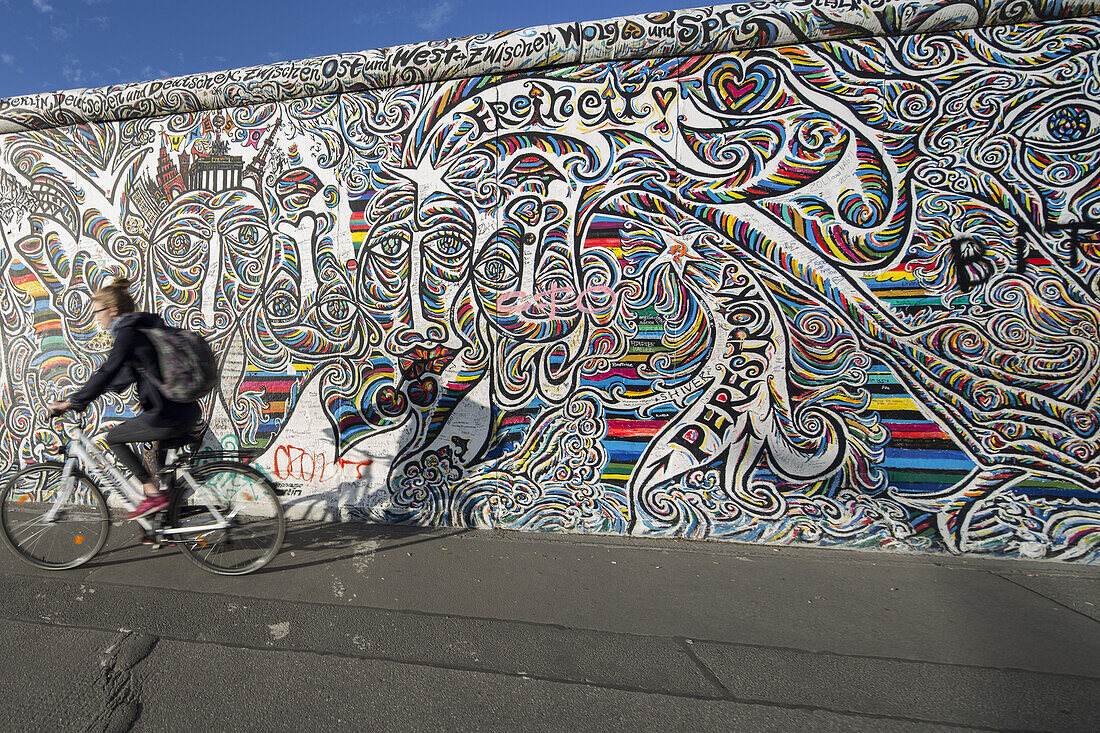 Frau fährt Fahrrad vorbei an der Berliner Mauer, East Side Gallery, Friedrichshain, Berlin, Deutschland