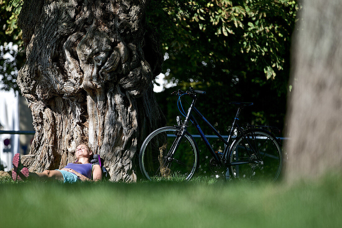 Junge Fahrradfahrerin ruht sich an einem großen Baum aus, Kempten, Bayern, Deutschland