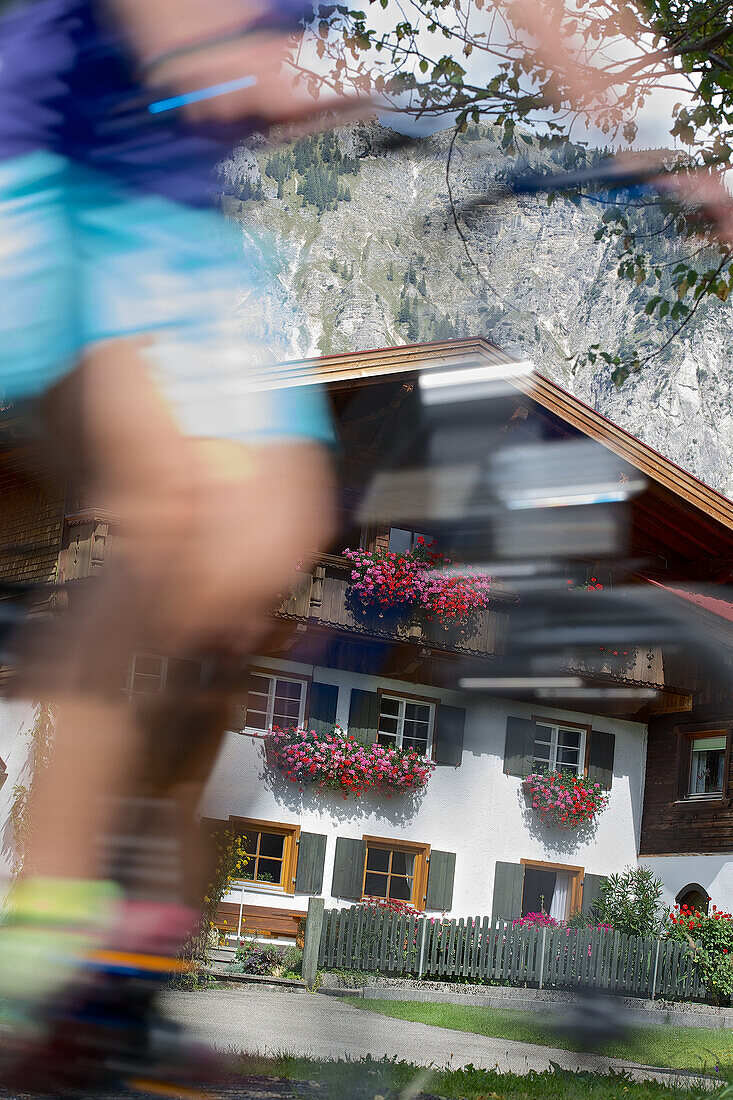 Young woman riding past a house on her bike on a sunny day, Tannheimer Tal, Tyrol, Austria